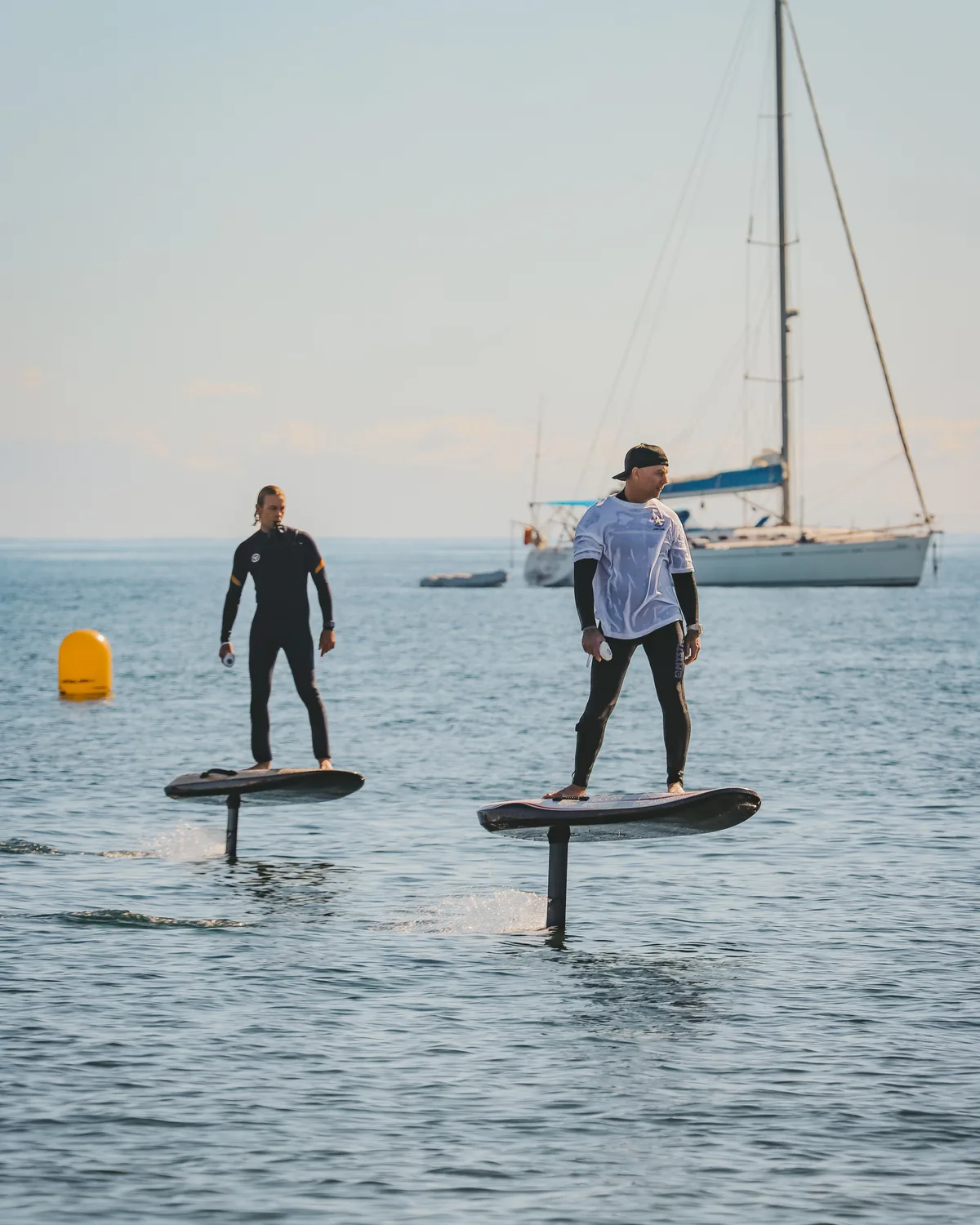 Dos riders de eFoil sobre aguas tranquilas con un velero al fondo